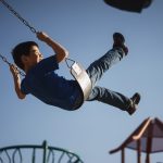 Child swinging high at playground under blue sky.