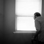 Elderly man looking out window with cane.