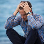 Man sitting, looking stressed by the water.